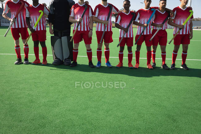 Front view low section of a multi-ethnic group of male field hockey players, preparing before a game, standing in a row on the pitch with their coach, holding hockey sticks on a sunny day — Stock Photo