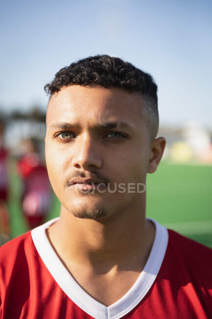 Portrait of a teenage mixed race male field hockey player, wearing a red team strip, standing on a field hockey pitch, looking to camera, focusing before a game, with his teammates in the background on a sunny day — Stock Photo