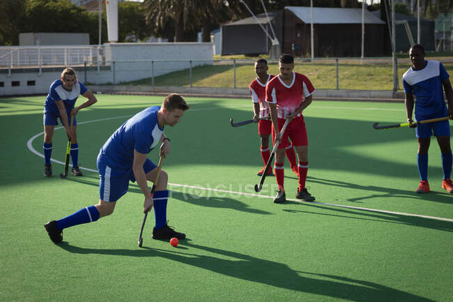 Side view of a teenage Caucasian male field hockey player, preparing to hit a ball on the pitch during a hockey game  between two multi-ethnic teenage male teams, on a sunny day — Stock Photo