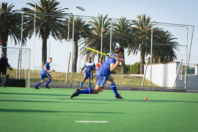 Side view of a teenage Caucasian male field hockey player preparing to hit a ball, during a hockey game between two multi-ethnic teenage male teams on a sunny day — Stock Photo