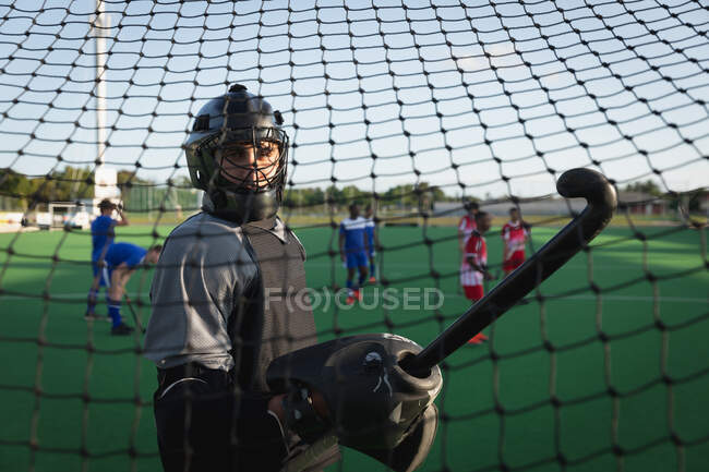 Vista lateral de un portero de hockey masculino caucásico, parado en la portería sosteniendo un palo de hockey, y girando para mirar a la cámara, durante un juego de hockey de campo en un día soleado, visto a través de la red de gol - foto de stock