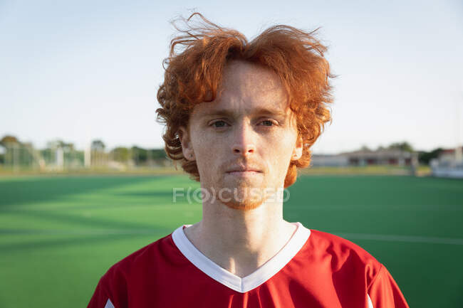 Portrait close up of a Caucasian male field hockey player with red hair, wearing a red team strip, standing on a hockey pitch looking to camera on a sunny day — Stock Photo