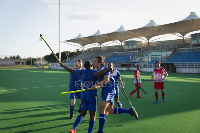 Side view of a multi-ethnic group of teenage male field hockey players, on the pitch, holding their hockey sticks and embracing, celebrating a victory on a sunny day — Stock Photo