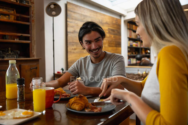 Вид спереди на молодого человека смешанной расы и молодую кавказскую женщину, сидящую за столом и наслаждающуюся завтраком вместе. — стоковое фото