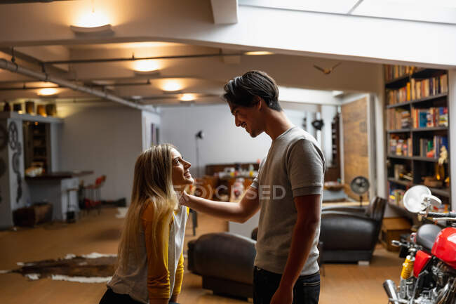 Vista lateral de uma jovem caucasiana e um jovem mestiço, aproveitando o tempo em casa, em pé em sua sala de estar, sorrindo e abraçando. — Fotografia de Stock