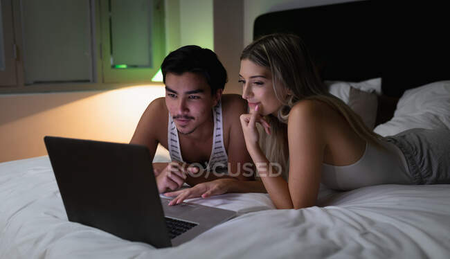 Front view of a young mixed race man and a young Caucasian woman enjoying time at home, lying in their bed and using their laptop computer. — Stock Photo