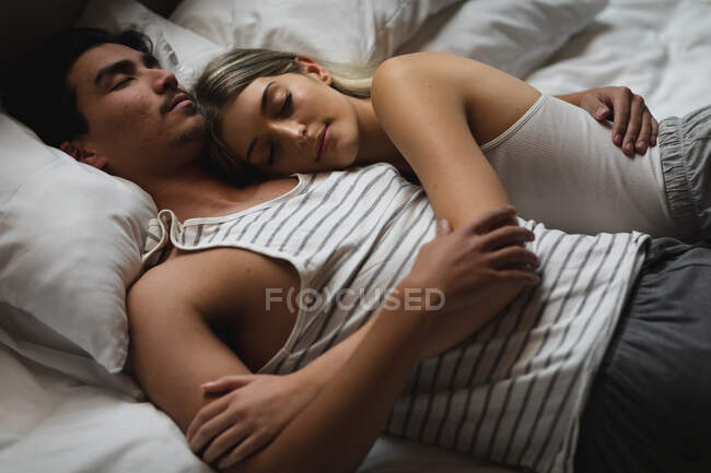 Вид сбоку на молодого человека смешанной расы и молодую кавказскую женщину, наслаждающуюся временем дома, спящую вместе, лежащую в своей постели и обнимающую. — стоковое фото