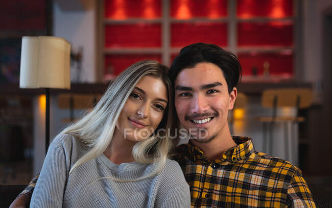 Portrait d'un jeune homme métis et d'une jeune femme caucasienne appréciant le temps passé à la maison, assise dans leur salon, souriante et regardant droit devant une caméra. — Photo de stock