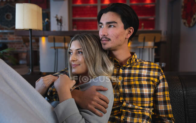 Vue de face gros plan d'un jeune homme métis et d'une jeune femme caucasienne profitant du temps passé à la maison, assis dans leur salon, embrassant et souriant. — Photo de stock
