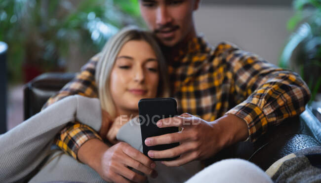 Вид спереди на молодого человека смешанной расы и молодую кавказку, наслаждающуюся временем дома, сидящую в гостиной, обнимающуюся и пользующуюся смартфоном. — стоковое фото