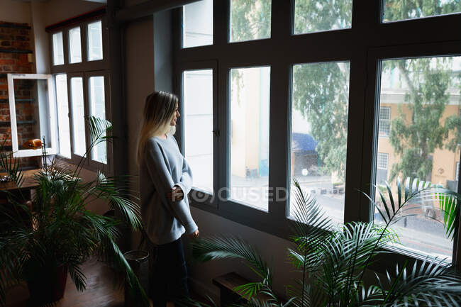 Selbstisolierung in Quarantäne. Seitenansicht einer jungen kaukasischen Frau, die die Zeit zu Hause genießt, einen grauen Pullover trägt, am Fenster steht und hindurchschaut. — Stockfoto