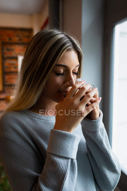 Front view close up of a young Caucasian woman enjoying time at home, wearing grey sweater, standing by the window and drinking coffee. — Stock Photo