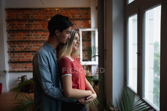 Вид сбоку на молодого человека смешанной расы и молодую кавказку, наслаждающуюся временем дома, стоящую у окна, обнимающую друг друга. — стоковое фото