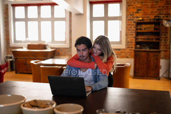 Vue de face d'une jeune femme caucasienne et d'un jeune homme de race mixte, profitant du temps passé à la maison, assis dans leur salon, souriant et embrassant tout en utilisant un ordinateur portable. — Photo de stock