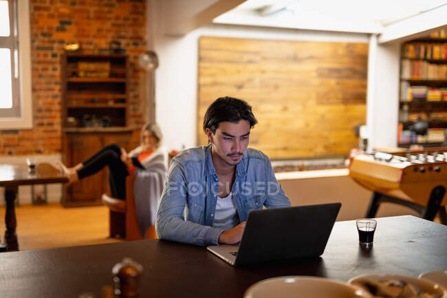 Vista frontal de un joven mestizo, disfrutando del tiempo en casa, sentado en la sala de estar, usando su computadora portátil con su pareja está sentado en el fondo. - foto de stock