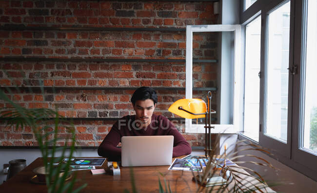 Selbstisolierung in Quarantäne. Frontansicht eines jungen Mannes mit gemischter Rasse, der in seinem Home Office sitzt und während der Arbeit seinen Laptop benutzt. — Stockfoto