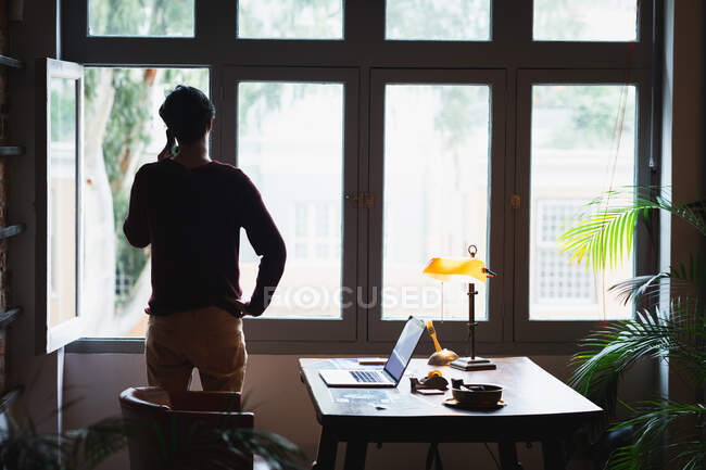 Vista trasera de un joven mestizo, parado en su oficina en casa junto a la ventana, hablando en su smartphone. - foto de stock