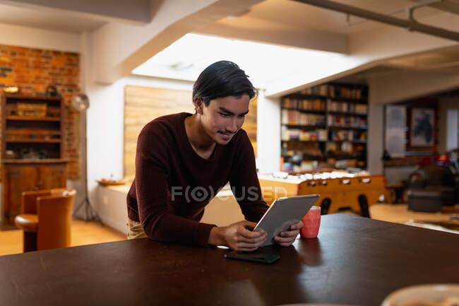 Frontansicht eines jungen Mannes mit gemischter Rasse, der Zeit zu Hause genießt, im Wohnzimmer steht, sein Tablet benutzt und lächelt. — Stockfoto