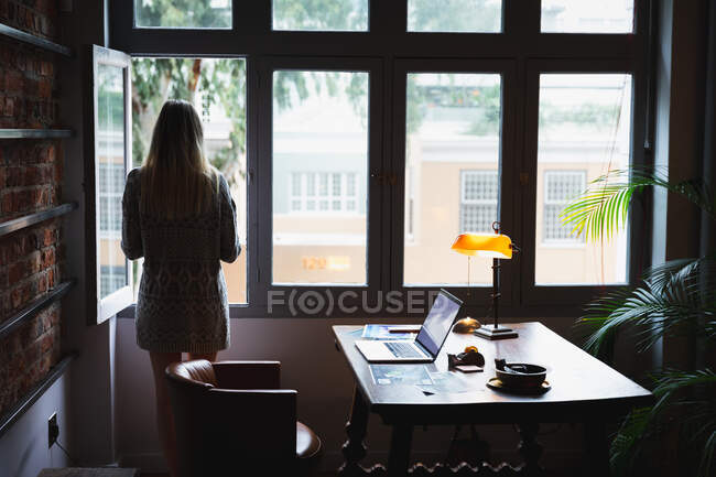 Vue arrière d'une jeune femme caucasienne, debout dans son bureau à la maison près de la fenêtre, faisant une pause tout en travaillant. — Photo de stock