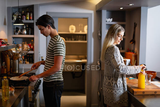 Vista lateral de um jovem homem de raça mista e uma jovem mulher caucasiana em pé na cozinha e preparando o café da manhã juntos . — Fotografia de Stock