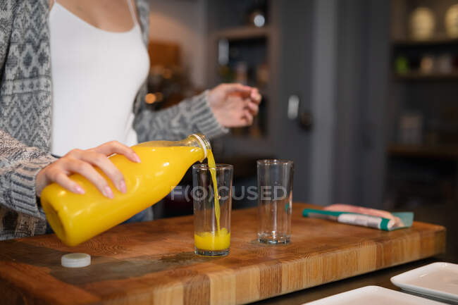 Vue de la partie médiane avant de la femme debout dans la cuisine préparant le petit déjeuner, versant du jus d'orange dans un verre. — Photo de stock