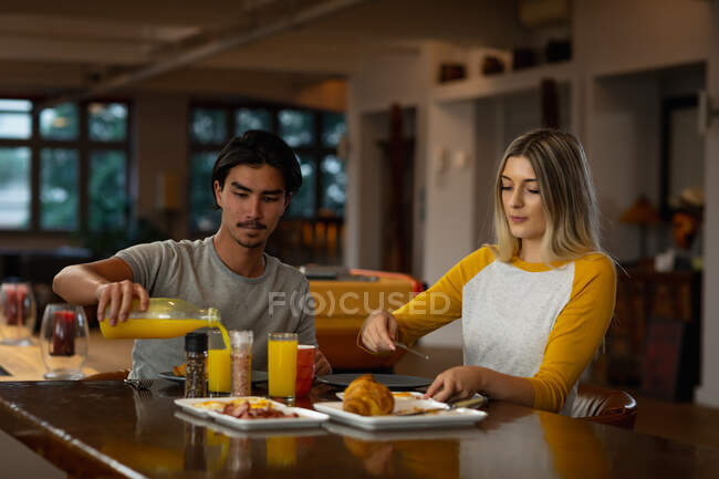 Vista frontal de un joven mestizo y una joven caucásica sentados junto a una mesa y disfrutando del desayuno juntos. - foto de stock