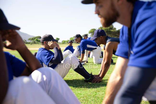 Vista lateral de cerca de un entrenador de béisbol masculino caucásico sosteniendo los pies de un jugador de béisbol de raza mixta, instruyendo a un grupo multiétnico de jugadores de béisbol masculino, entrenando antes de un juego, haciendo ejercicio sentado en un campo de juego en un día soleado - foto de stock