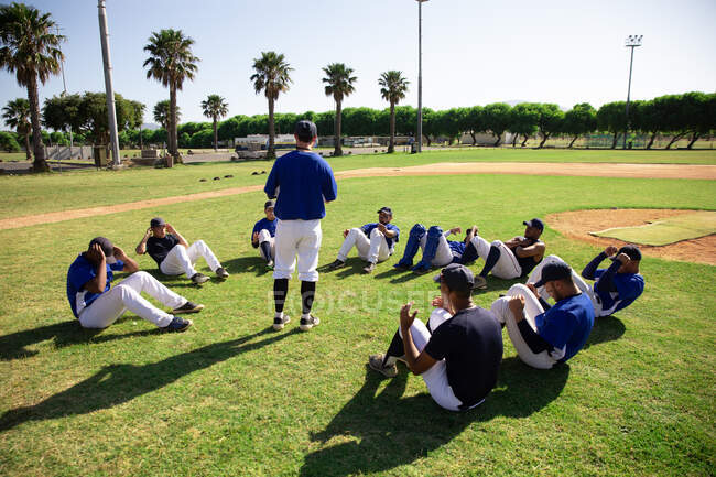 Vista lateral de un grupo multiétnico de jugadores de béisbol masculinos sosteniéndose los pies unos a otros, entrenándose antes de un juego, ejercitándose haciendo sentadas en un círculo, en un campo de juego en un día soleado - foto de stock