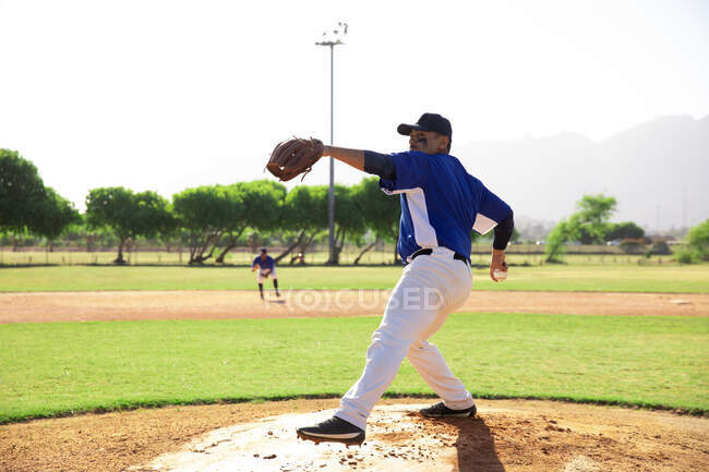 Vista laterale di un giocatore di baseball maschile di razza mista, che lancia una palla durante una partita di baseball, in una giornata di sole — Foto stock