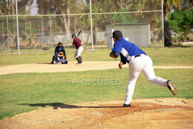 Вид ззаду змішаної раси чоловічий бейсболіст під час бейсбольної гри, штовхаючи м'яч, з гіркотою і ловцем на задньому плані в сонячний день — стокове фото