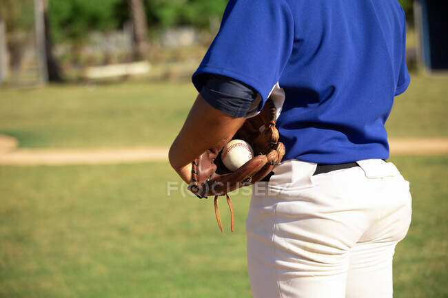 Vista lateral de la sección media del jugador de béisbol masculino, sosteniendo una pelota en su guante durante un juego de béisbol, en un día soleado - foto de stock