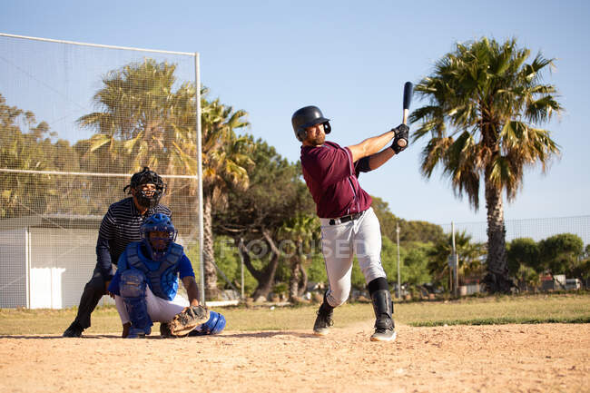 Вид спереди на белого бейсболиста во время игры в бейсбол в солнечный день, удар по мячу бейсбольной битой, кэтчер и другой игрок сидят на корточках за киллером — стоковое фото