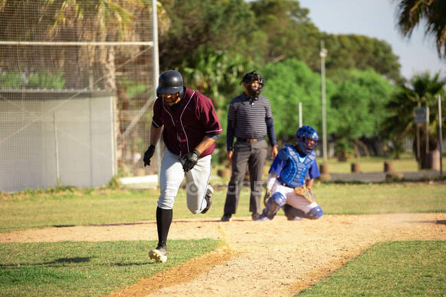 Front view of a Caucasian male baseball player during a baseball game on a sunny day, running after hitting a ball with a baseball bat, with a catcher and another player in the background — Stock Photo