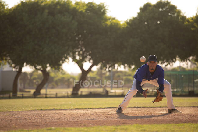 Передній вигляд змішаної раси чоловічий бейсболіст під час бейсбольної гри в сонячний день, готуючись зловити низький м'яч у рукавичці — стокове фото