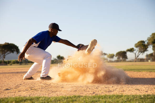 Vista lateral de un jugador de béisbol masculino de raza mixta, durante un partido de béisbol en un día soleado, sosteniendo una pelota en su guante, tocando a un oponente que hizo una nube de arena - foto de stock