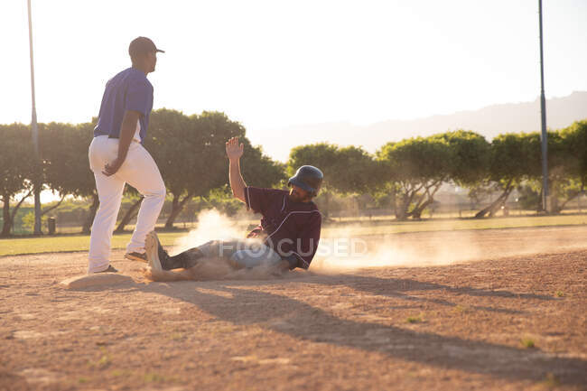 Вид сбоку на белого бейсболиста во время игры в бейсбол в солнечный день, сидящего рядом с базой, разочарованного, когда его противник уходит — стоковое фото
