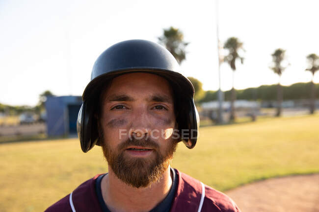 Retrato de un jugador de béisbol caucásico, con uniforme de equipo y casco, parado en un campo de béisbol, mirando a una cámara - foto de stock
