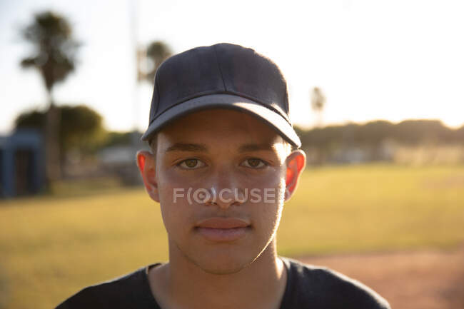 Retrato de un jugador de béisbol masculino de raza mixta, usando un uniforme de equipo y una gorra, parado en un campo de béisbol, mirando a una cámara - foto de stock