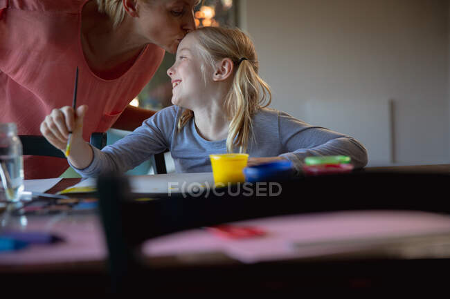 Вид спереди на кавказку, наслаждающуюся семейным отдыхом с дочерью дома, сидящую за столом в гостиной, рисующую и улыбающуюся, дающую ей поцелуй — стоковое фото