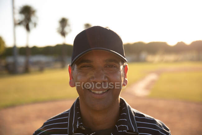 Портрет змішаної раси чоловічий бейсбольний арбітр, одягнений в уніформу і кепку, стоїть на бейсбольному полі, дивлячись на камеру, посміхаючись — стокове фото
