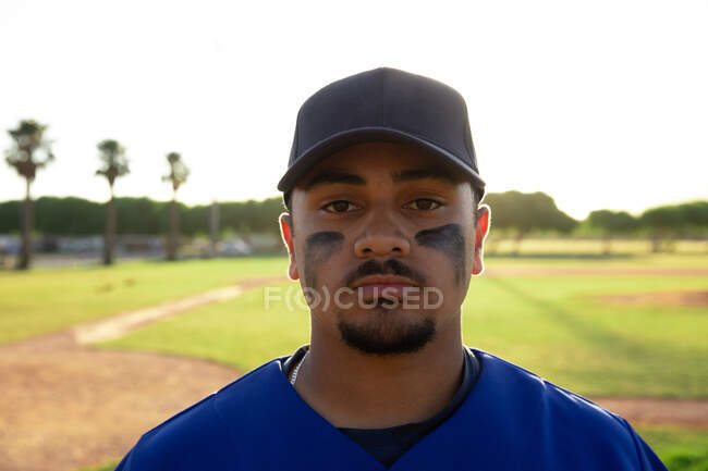 Portrait of a mixed race male baseball player, wearing a team uniform and a cap, standing on a baseball field, looking at camera — Stock Photo