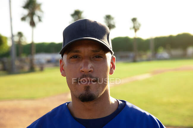 Портрет змішаної раси чоловіка бейсболіста, одягнений в командну форму і кепку, стоїть на бейсбольному полі, дивлячись на камеру — стокове фото