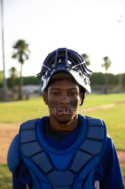 Retrato de un jugador de béisbol masculino de raza mixta, con uniforme de equipo, casco y almohadillas en el pecho, de pie en un campo de béisbol, mirando a la cámara - foto de stock