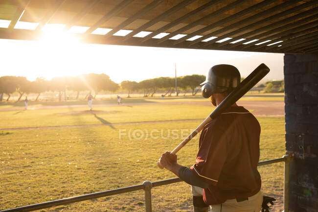 Visão traseira de um jogador de beisebol masculino caucasiano, assistindo a um jogo de beisebol atrás de uma barreira, descansando um taco de beisebol em seu ombro, em um dia ensolarado — Fotografia de Stock