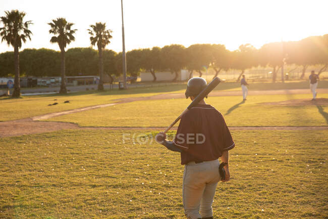 Vista trasera de un jugador de béisbol caucásico, mirando un partido de béisbol, apoyando un bate de béisbol en su hombro, caminando hacia un campo de béisbol, en un día soleado - foto de stock