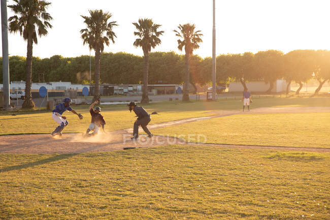 Vista lateral de tres jugadores de béisbol masculino de raza mixta durante un juego de béisbol en un día soleado, uno se desliza hacia una base, y uno está tratando de tocarlo con una pelota - foto de stock