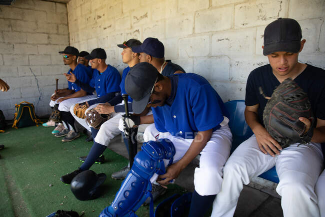 Вид збоку на ряд багатоетнічних чоловічих бейсболістів, які готуються перед грою, сидячи в роздягальні, одягаючи ноги, фокусуючись, поки вони чекають, взаємодіючи — стокове фото