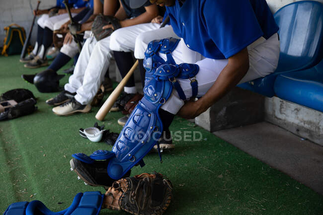 Sección baja vista lateral del jugador de béisbol masculino preparándose antes de un partido, sentado en un vestuario, poniéndose sus almohadillas para las piernas, con sus compañeros de equipo sentados en una fila en el fondo - foto de stock
