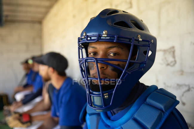 Portrait of a mixed race male baseball player, preparing before a game, wearing a team uniform, a helmet and chest pads, sitting in a changing room, looking at a camera, with his teammates in the background — Stock Photo