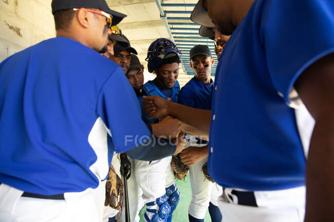 Side view of a multi-ethnic group of male baseball players, preparing before a game, standing in a changing room, team huddling, fist bumping, interacting, motivating each other — Stock Photo